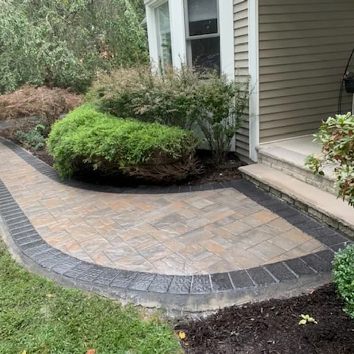 Curved stone paver walkway with dark border beside a house and landscaped shrubs.