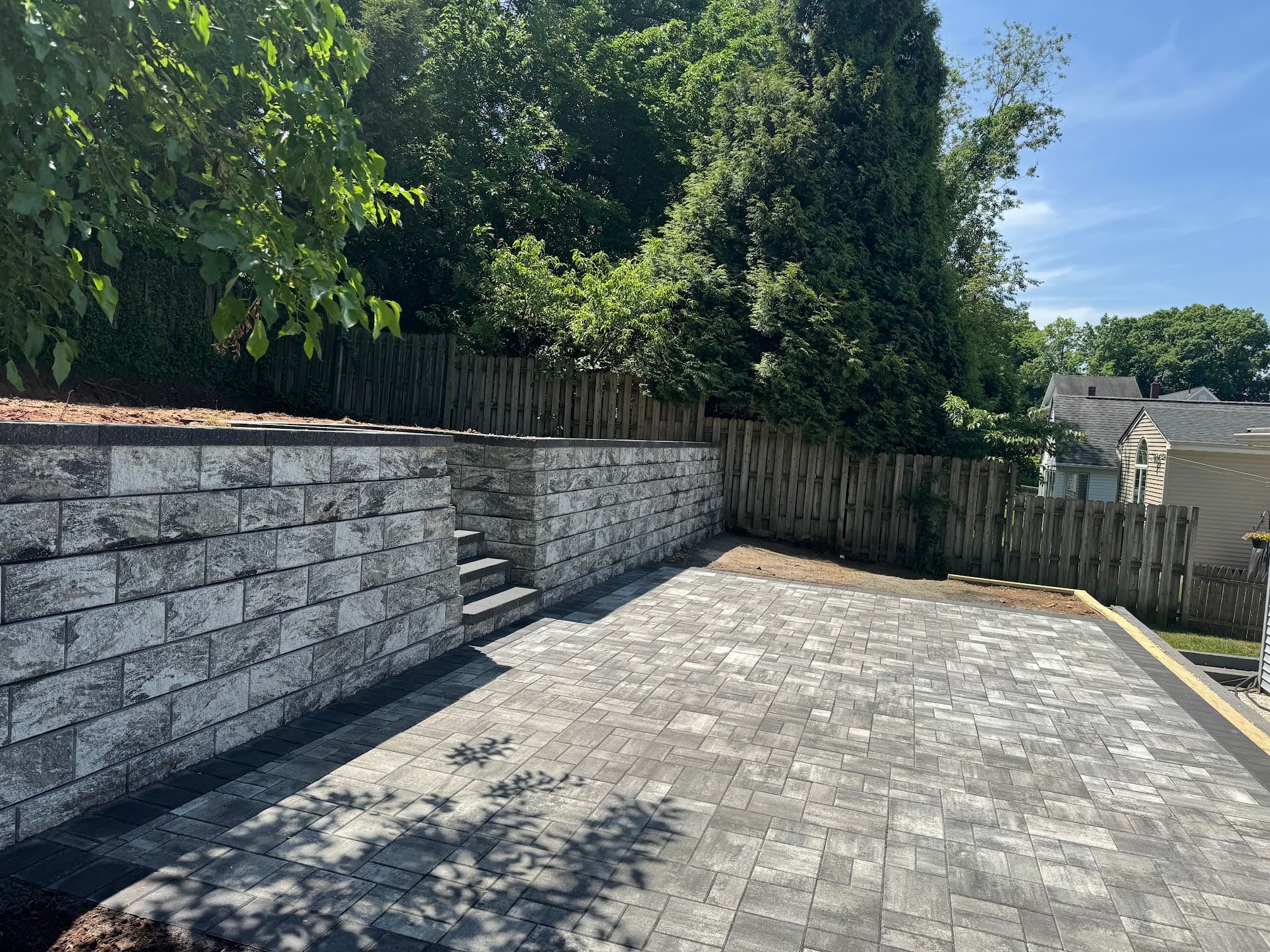 Gray stone retaining wall with steps beside a newly installed paver patio in a backyard.